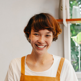 Smiling woman with short brown hair, wearing a white shirt and orange apron, indoors beside a window.