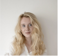 Portrait of a blonde woman with wavy hair wearing a white blouse, against a plain light background.