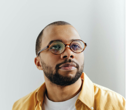 Portrait of a man with short dark hair and a beard, wearing round glasses and a yellow shirt against a light background.